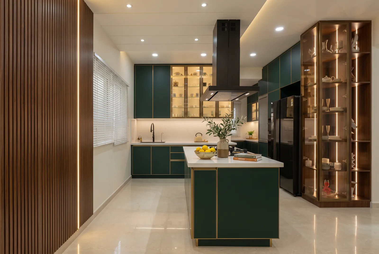 An island kitchen with bottle-green matt shutters, brass edges, a fluted oak wall and a lit bronze display tower.