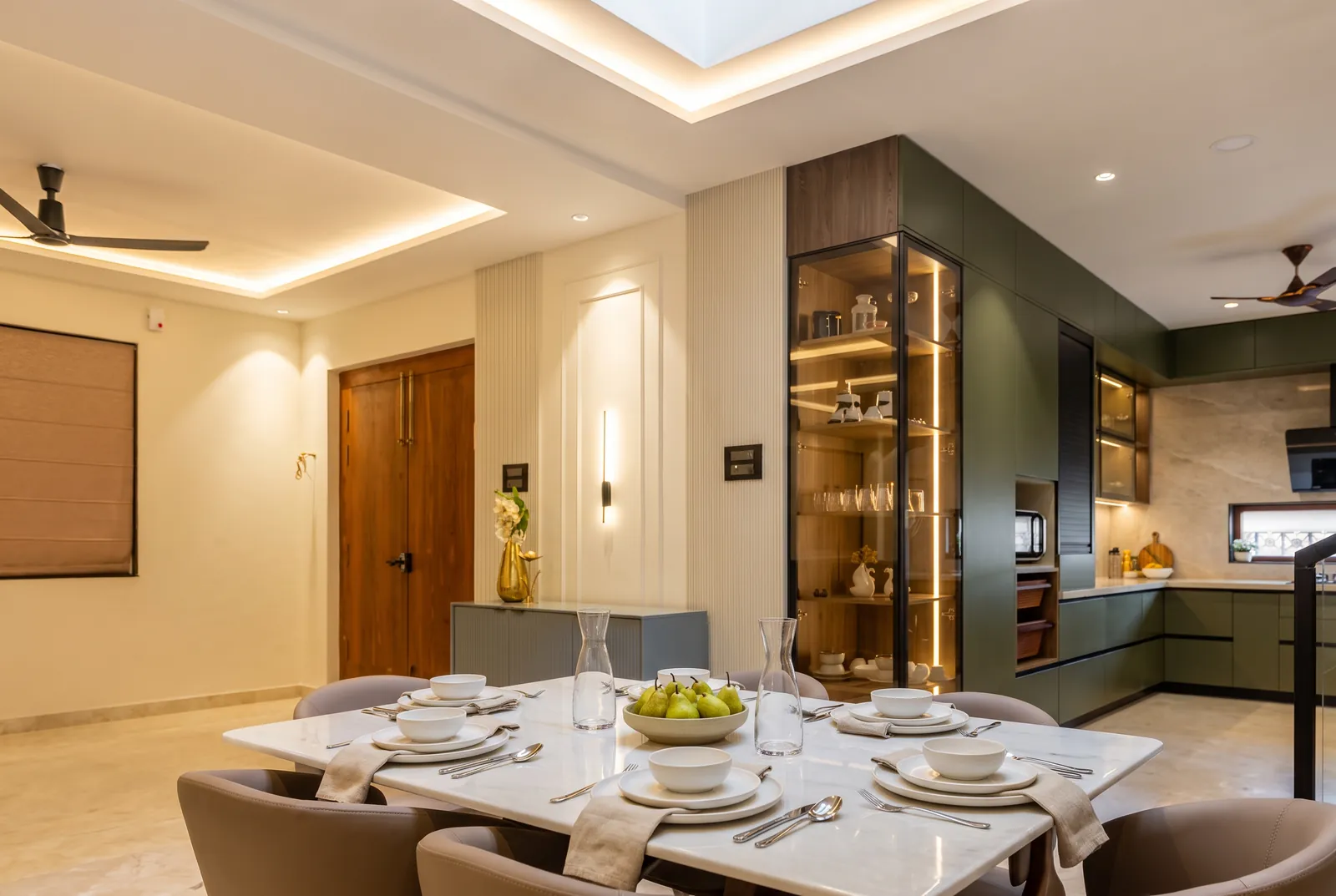 A dining room with a lit smoked-glass crockery unit and an olive-green kitchen beyond.