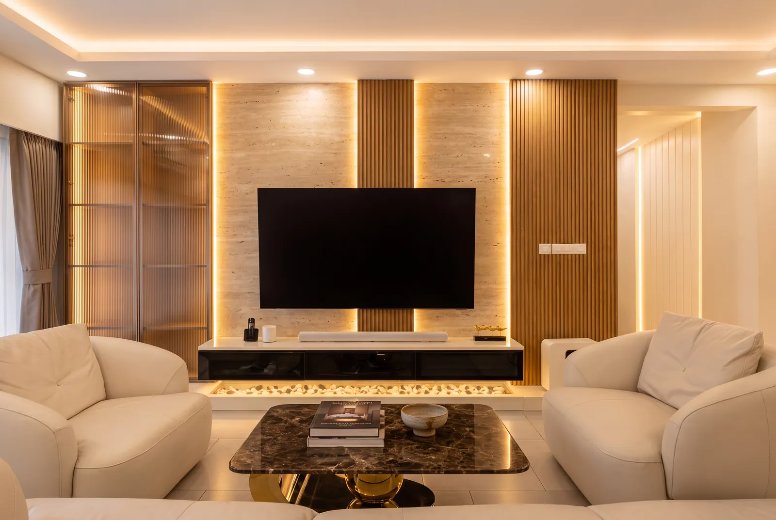 A living room TV wall in travertine and fluted oak, with a backlit bronze-glass cabinet and curved cream sofas.
