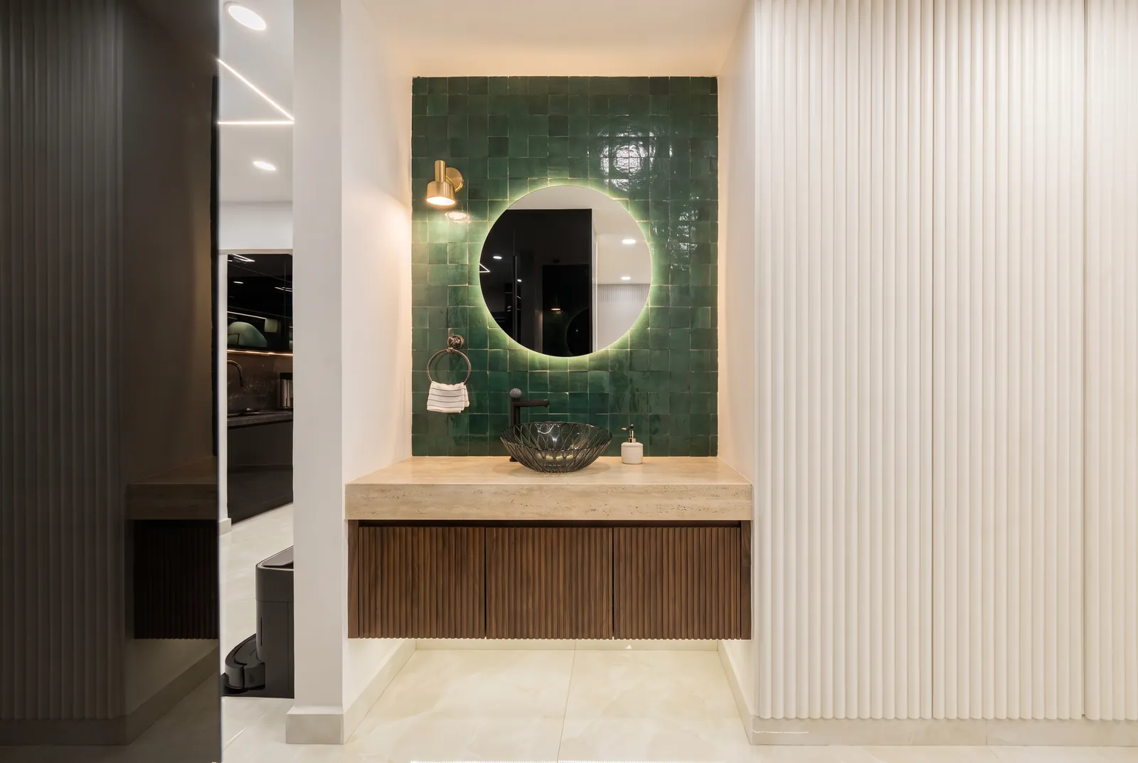 A vanity with green zellige tiles, a round backlit mirror, a travertine top and a fluted walnut cabinet.